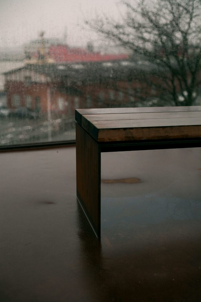 A sleek bench sits indoors against a rainy window, reflecting a muted urban scene outside.