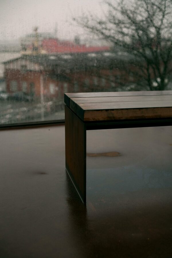 A sleek bench sits indoors against a rainy window, reflecting a muted urban scene outside.